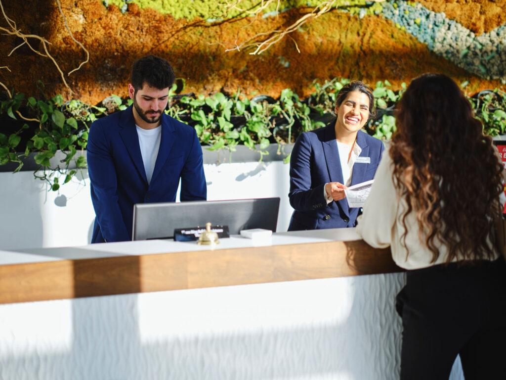 Two receptionists assisting a woman at a modern, plant-decorated front desk.