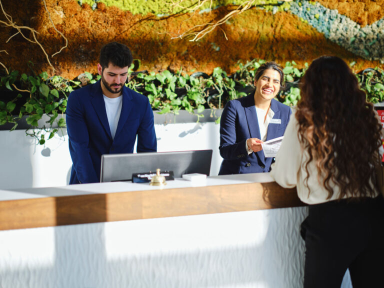 Two receptionists assisting a woman at a modern, plant-decorated front desk.